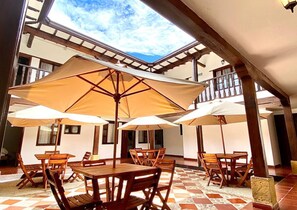 An outdoor patio with wooden tables and chairs, covered by large umbrellas, and a view of the sky through a skylight. - Hotel Calle Real (Villa de Leyva)