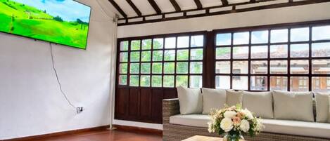 Una habitación con televisor montado en la pared, un sofá con cojines, una mesa de centro con un jarrón de flores y ventanas amplias con vistas a zonas verdes y edificios.