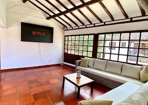 A living room with a TV mounted on the wall, a sofa, a coffee table, and a large window with a view of buildings. - Hotel Calle Real (Villa de Leyva)