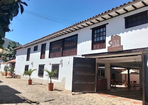 A white building with a tiled roof, a sign reading 'Hotel Cuca Real,' and a covered entrance with chairs and tables. - Hotel Calle Real (Villa de Leyva)