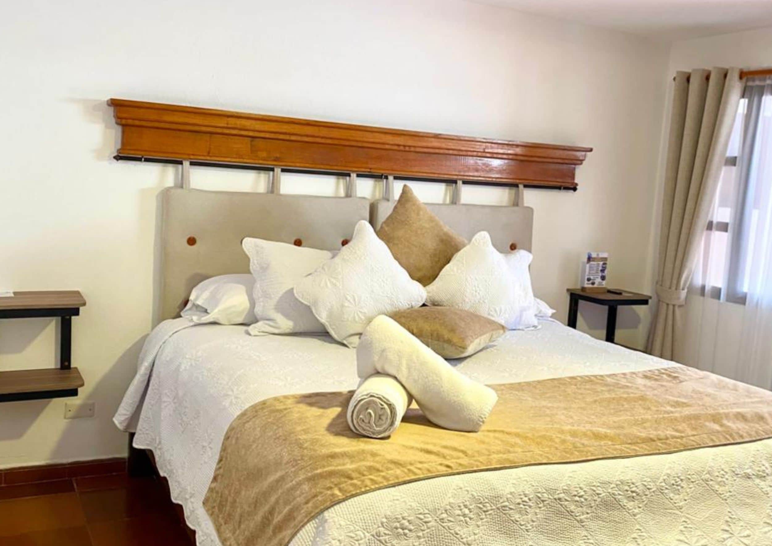 Una cama bien tendida con cabecera de madera, varios almohadones y una toalla enrollada.