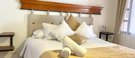 Una cama bien tendida con cabecera de madera, varios almohadones y una toalla enrollada.