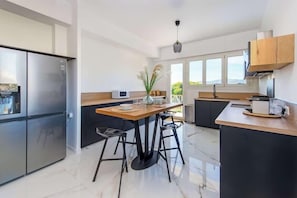 A modern kitchen with a wooden dining table, black stools, a stainless steel refrigerator, and a large window providing natural light. - Villa App Grenouillere (Cagnes-sur-Mer)