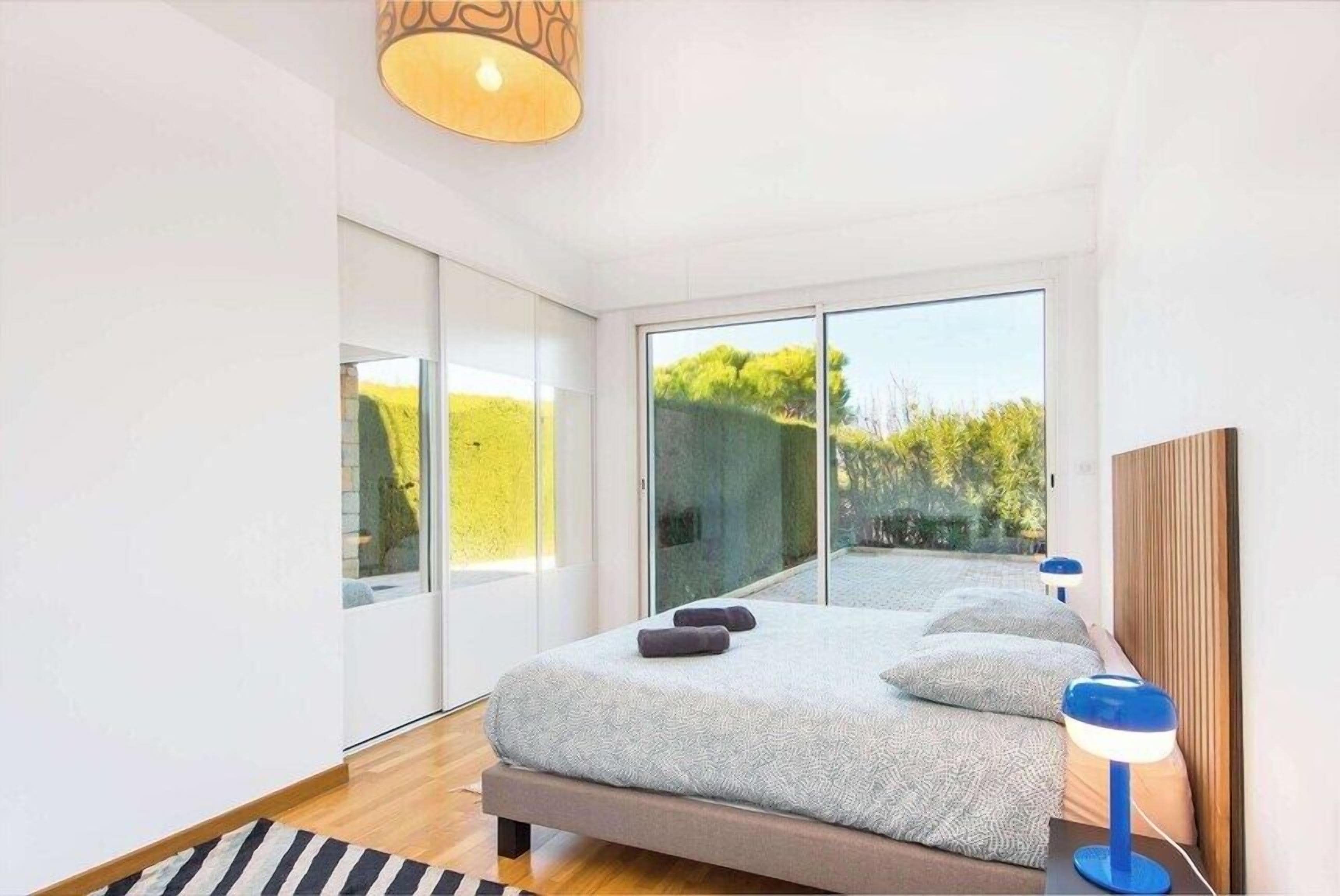 A modern bedroom with a large bed, sliding glass doors, and a view of greenery.