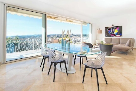 A modern living room with a glass dining table, four chairs, a sofa, and a large window offering a city view.