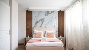 A bedroom with a bed, two nightstands, a headboard with a marble-like design, wooden panels, and a window with sheer curtains. - Villa Alexandre Dumas (Bussy-Saint-Georges)