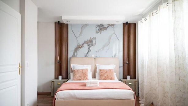 Een slaapkamer met een bed, twee nachtkastjes, een hoofdbord met een marmerachtig patroon, houten lambrisering en een raam met dunne gordijnen.
