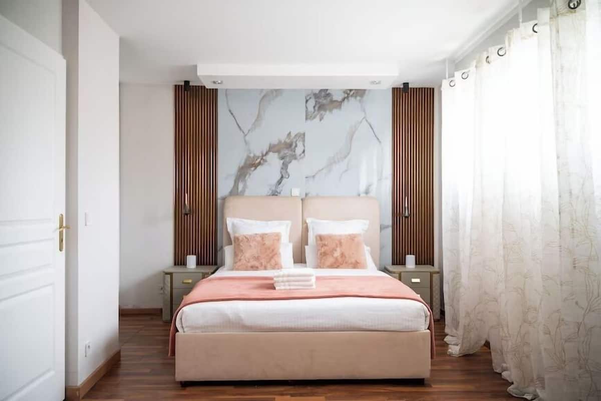 A bedroom with a bed, two nightstands, a headboard with a marble-like design, wooden panels, and a window with sheer curtains.