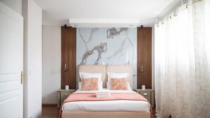 A bedroom with a bed, two nightstands, a headboard with a marble-like design, wooden panels, and a window with sheer curtains. - Villa Alexandre Dumas (Bussy-Saint-Georges)