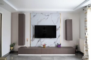 Una sala de estar moderna con un televisor de pantalla plana grande montado en una pared de mármol, un mueble de entretenimiento de color oscuro debajo y una planta pequeña en el suelo.