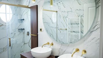 Een moderne badkamer met een grote ronde spiegel, twee wastafels met gouden kranen, een douchecabine van glas en marmeren wanden.