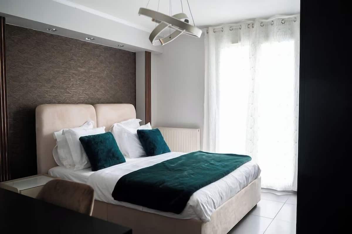 A modern hotel room with a large bed, a headboard with a textured design, a ceiling light fixture, and a window with sheer curtains.