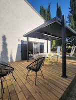 Una terraza de madera con dos sillas negras y una mesa, un área de patio techada y un edificio blanco con una puerta y ventanas.