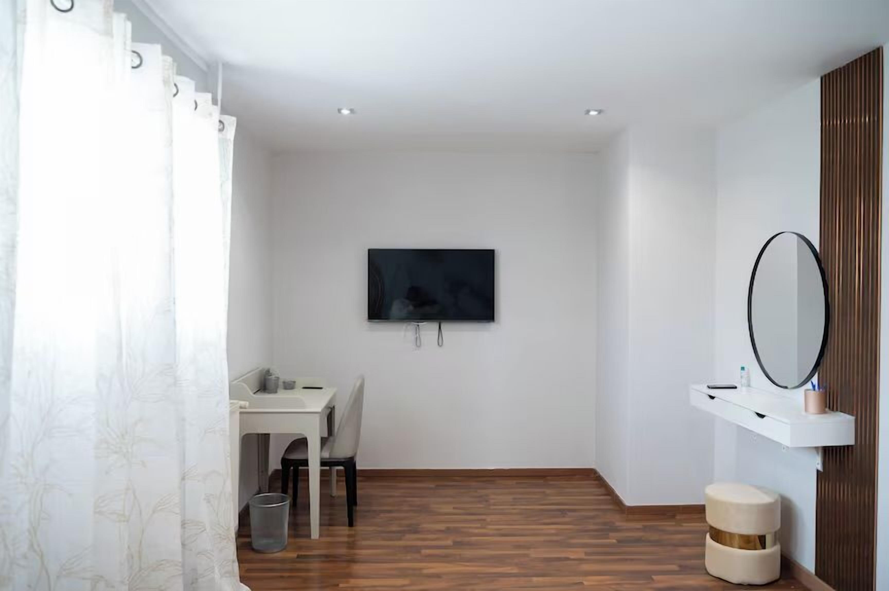 A modern room with a wooden floor, a white desk with a chair, a TV mounted on the wall, a small round stool, and a mirror on a white shelf.