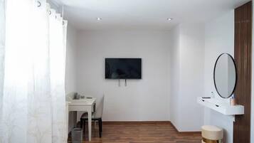 A modern room with a wooden floor, a white desk with a chair, a TV mounted on the wall, a small round stool, and a mirror on a white shelf.