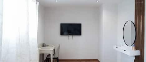 Una habitación moderna con suelo de madera, un escritorio blanco con silla, televisor montado en la pared, un taburete pequeño y redondo, y un espejo sobre una estantería blanca.