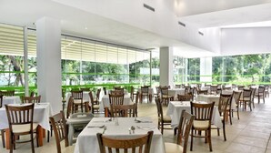 A restaurant with multiple tables and chairs, large windows with a view of greenery, and a modern interior design. - Villas Holidays (Kiten)