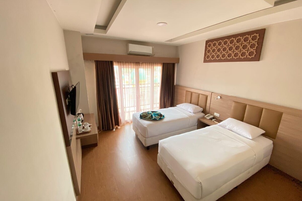 A hotel room with two beds, a wooden headboard, a flat-screen TV, a small table with a phone and a bottle, and a window with curtains.