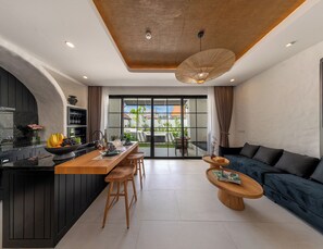 A modern kitchen with a central island, a dining area with a wooden table and chairs, and a living area with a dark blue sofa and a wooden coffee table. - Kiara Ocean Place (Tibubeneng)