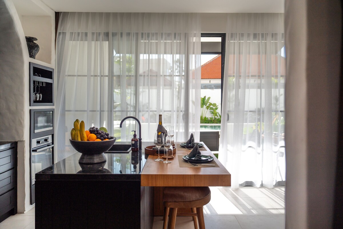 A modern kitchen with a central island, a bowl of fruit, and a dining area with chairs and a table set for two.