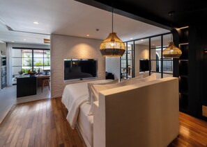 A modern bedroom with a large bed, a TV, and a dining area with a view of greenery. - Kiara Ocean Place (Tibubeneng)