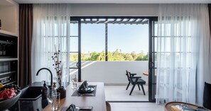 A modern kitchen with a dining area, a balcony with a table and chairs, and a view of greenery. - Kiara Ocean Place (Tibubeneng)