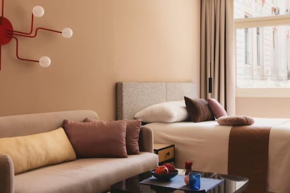 A hotel room with a sofa, a bed with pillows, a small table with a bowl and a vase, and a window with curtains. - Saint J Hotel By Brassbell (Cairo)