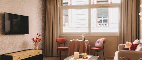 Un salon moderne avec un téléviseur à écran plat, une table basse en bois, deux fauteuils, un canapé et une petite table ornée de bougies et de livres.
