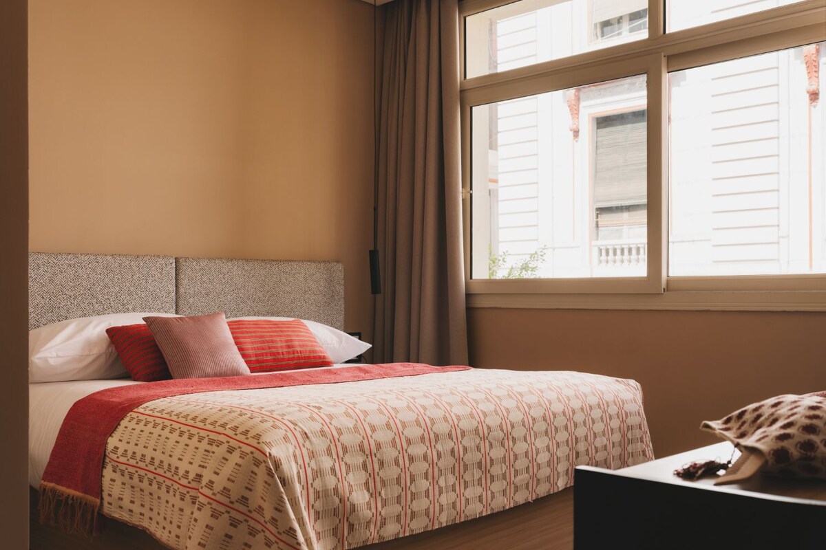 Un dormitorio con cama, mesita de noche, ventana con cortinas y vistas a edificios.