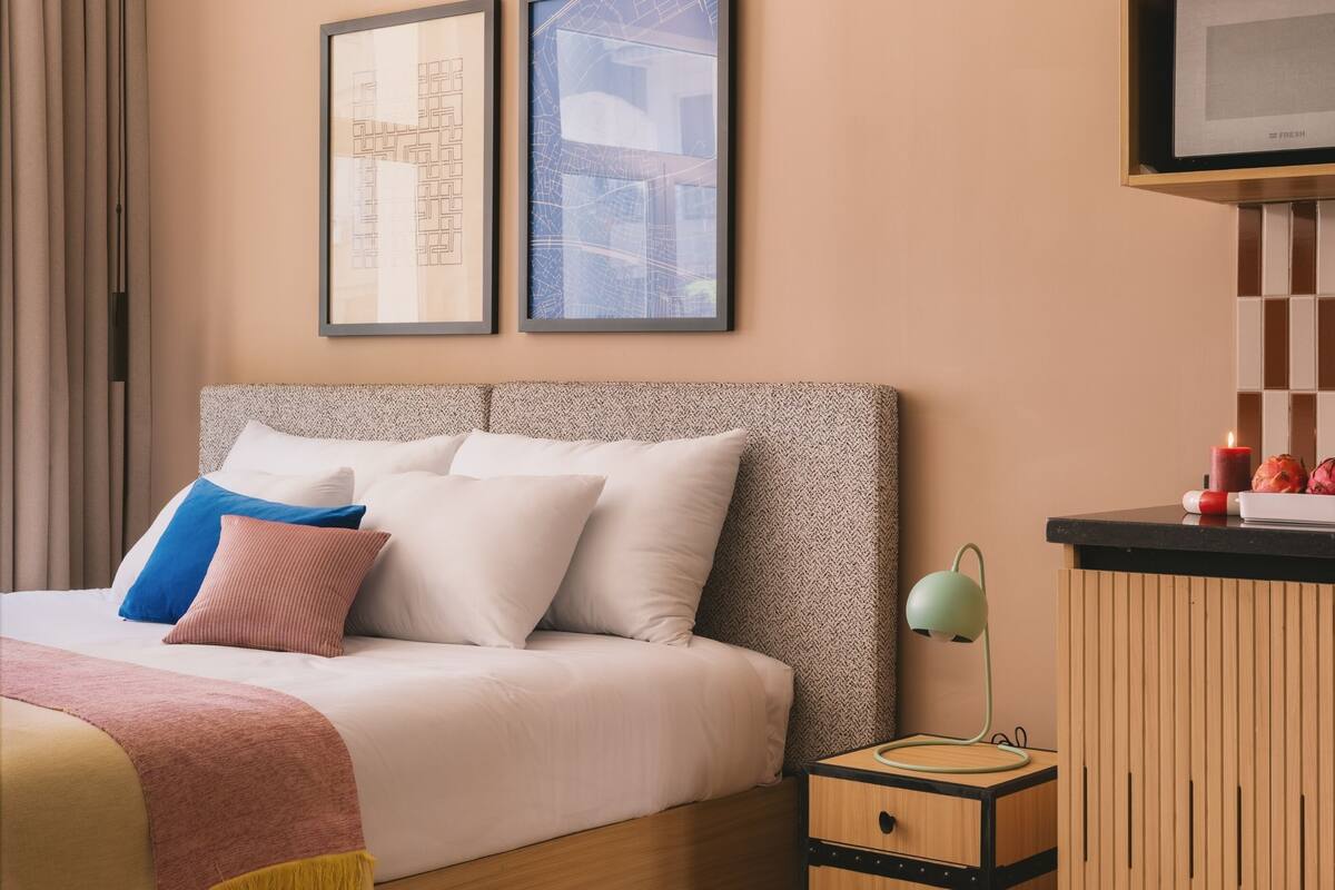 Una habitación de hotel ordenada con una cama, una mesita de noche con lámpara y cuadros en la pared.