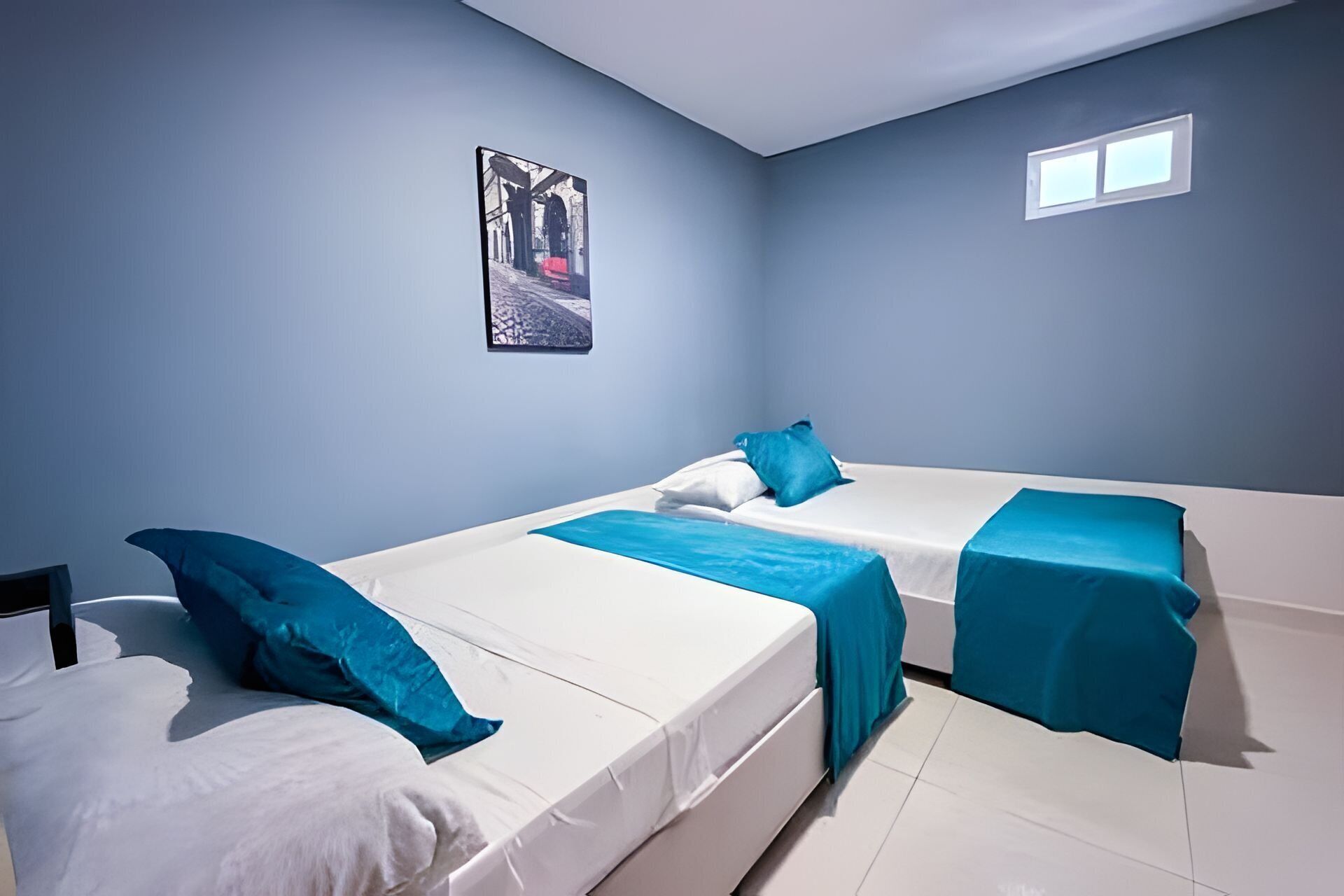 A room with two beds, each with white bedding and blue accents. There is a small window on the wall and a framed picture above the beds.