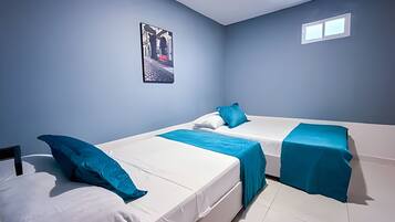 A room with two beds, each with white bedding and blue accents. There is a small window on the wall and a framed picture above the beds.
