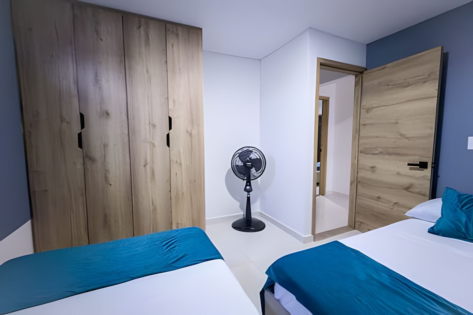 A bedroom with two beds, a wooden wardrobe, a standing fan, and a door leading to another room.