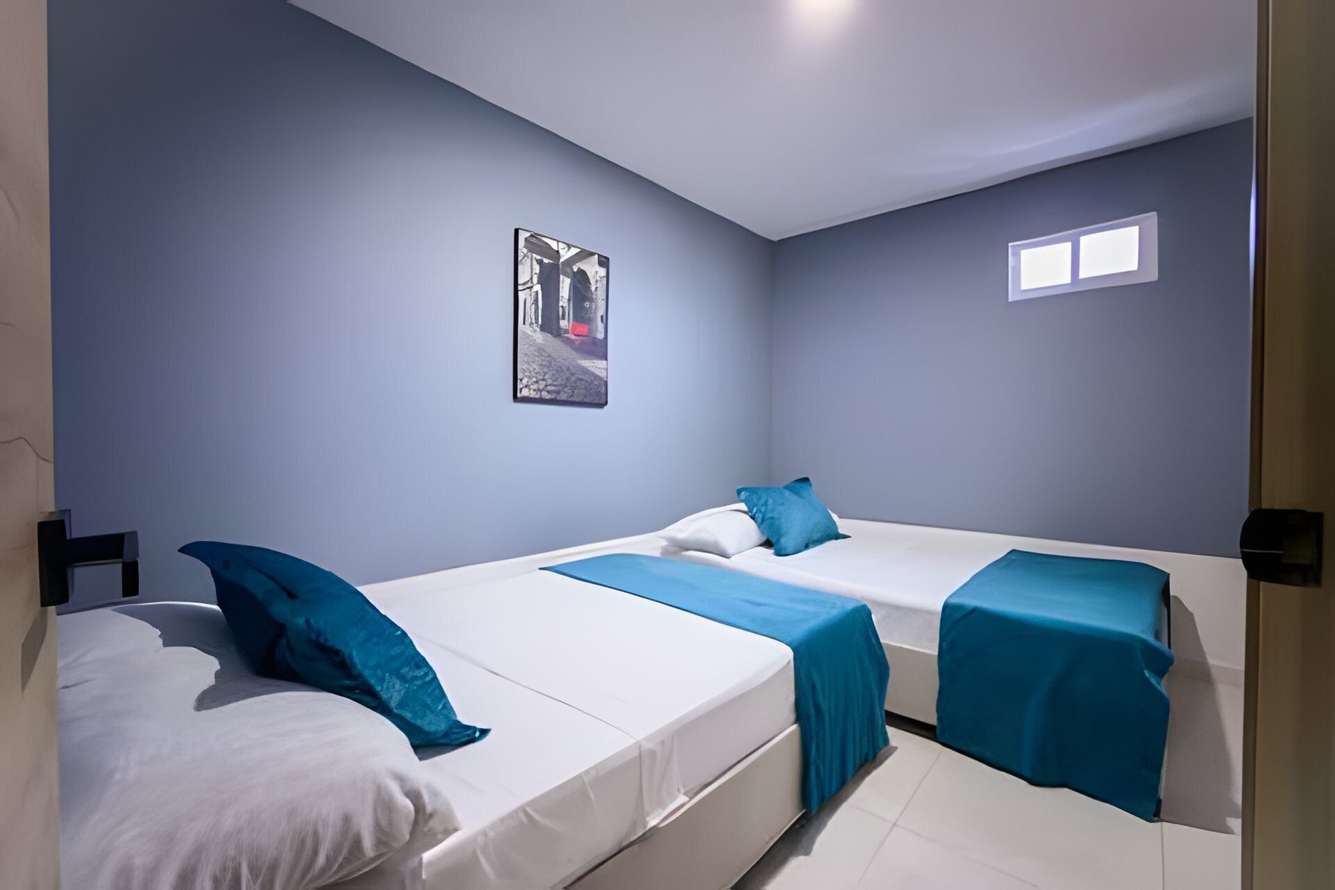 A room with three beds, each with a blue bedspread and pillows, and a framed picture on the wall.