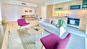 A modern living room with a white sofa, two purple chairs, a small round table, and a kitchen area with wooden cabinets and a microwave. - Nice 2Bdrm Unit - Downtown Miami (Miami)
