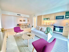 A modern living room with a white sofa, two purple chairs, a small round table, and a kitchen area with wooden cabinets and a microwave. - Nice 2Bdrm Unit - Downtown Miami (Miami)