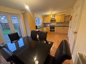 A modern kitchen with wooden cabinets, a black dining table with four chairs, and a view of the backyard through large sliding glass doors. - 4br- Garden- Parking- WiFi- Near uea and nnuh (Norwich)