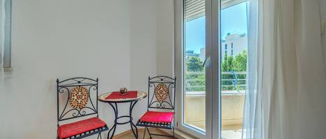 A room with two metal chairs and a small table, a sliding glass door to an outdoor balcony, and a view of buildings and greenery outside.