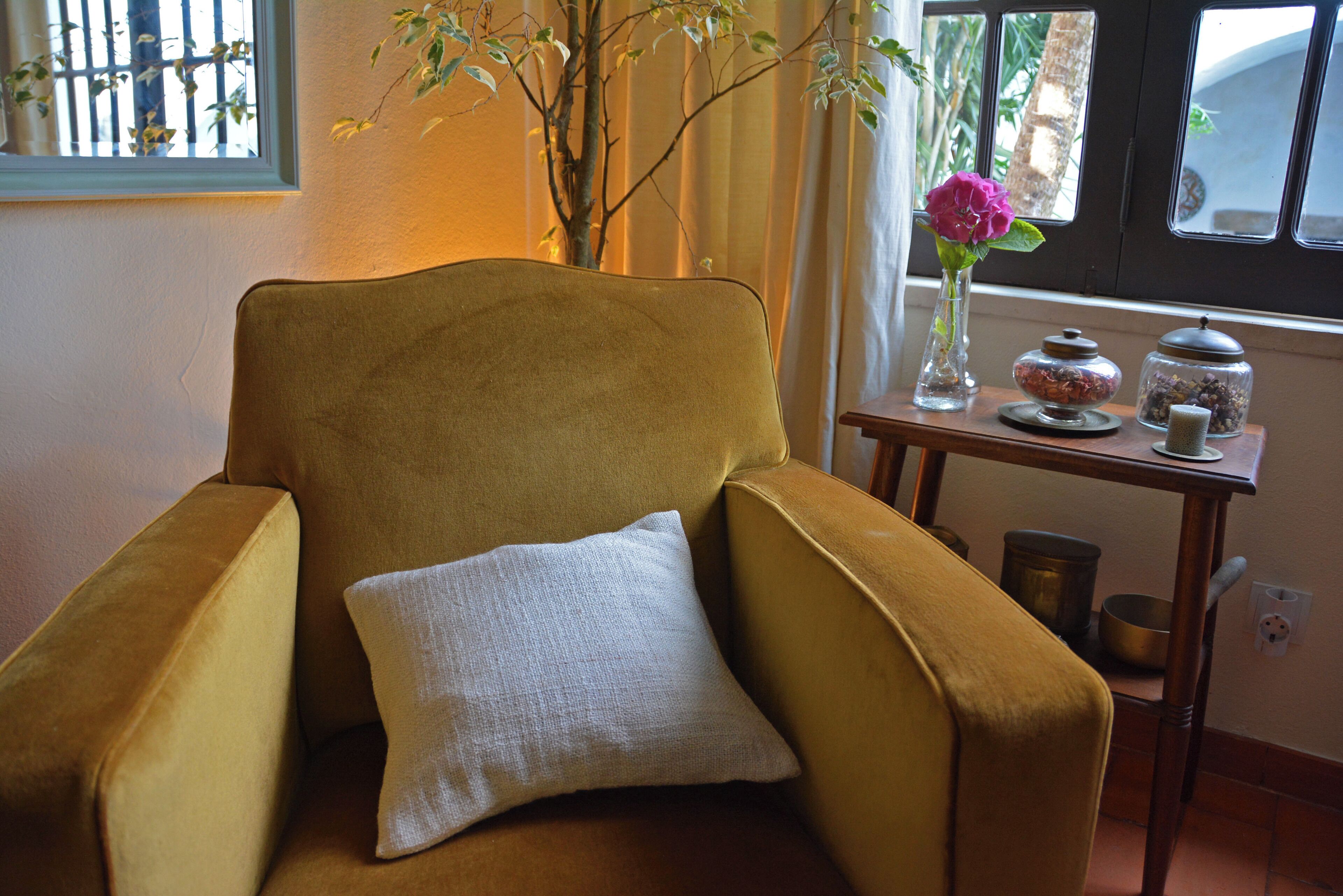 A mustard-colored armchair with a white pillow, a vase with a pink flower, and a small wooden table with jars and a bowl.