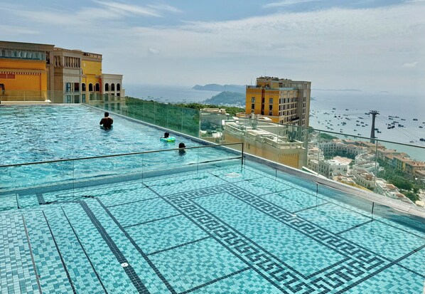 A rooftop infinity pool with clear blue water, surrounded by buildings with balconies overlooking the sea.