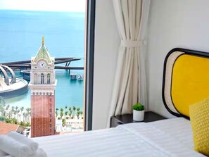 A hotel room with a bed, a small potted plant, and a view of a clock tower and water body.