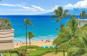 A beach view with palm trees, a sandy beach, and a clear blue ocean. - Maui Resort Rentals: The Whaler 720/4 - 7th Floor 2BR with Gorgeous Partial Ocean and West Maui Mountain Views! (Lahaina)
