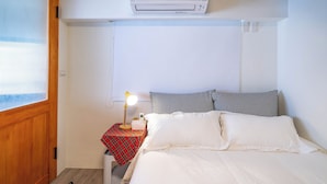 Una habitación con una cama, una mesita de noche con lámpara, aire acondicionado instalado en la pared y una ventana con persianas.