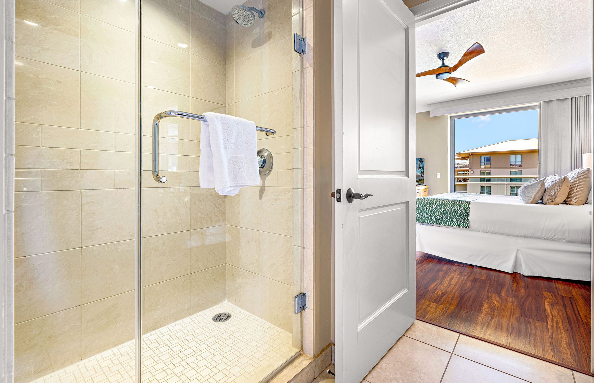Une chambre d’hôtel avec une douche en verre, une porte blanche, un lit recouvert d’un couvre-lit à motifs verts, et une vue sur un bâtiment à l’extérieur.