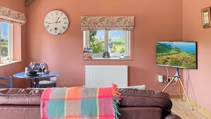 Um quarto acolhedor com um sofá de couro marrom, uma mesa redonda com toalha de mesa azul, um relógio, uma janela com vista e uma televisão sobre um tripé.