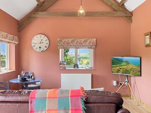 Ein gemütliches Zimmer mit einem braunen Ledersofa, einem runden Tisch mit blauer Tischdecke, einer Uhr, einem Fenster mit Aussicht und einem Fernseher auf einem Stativ.