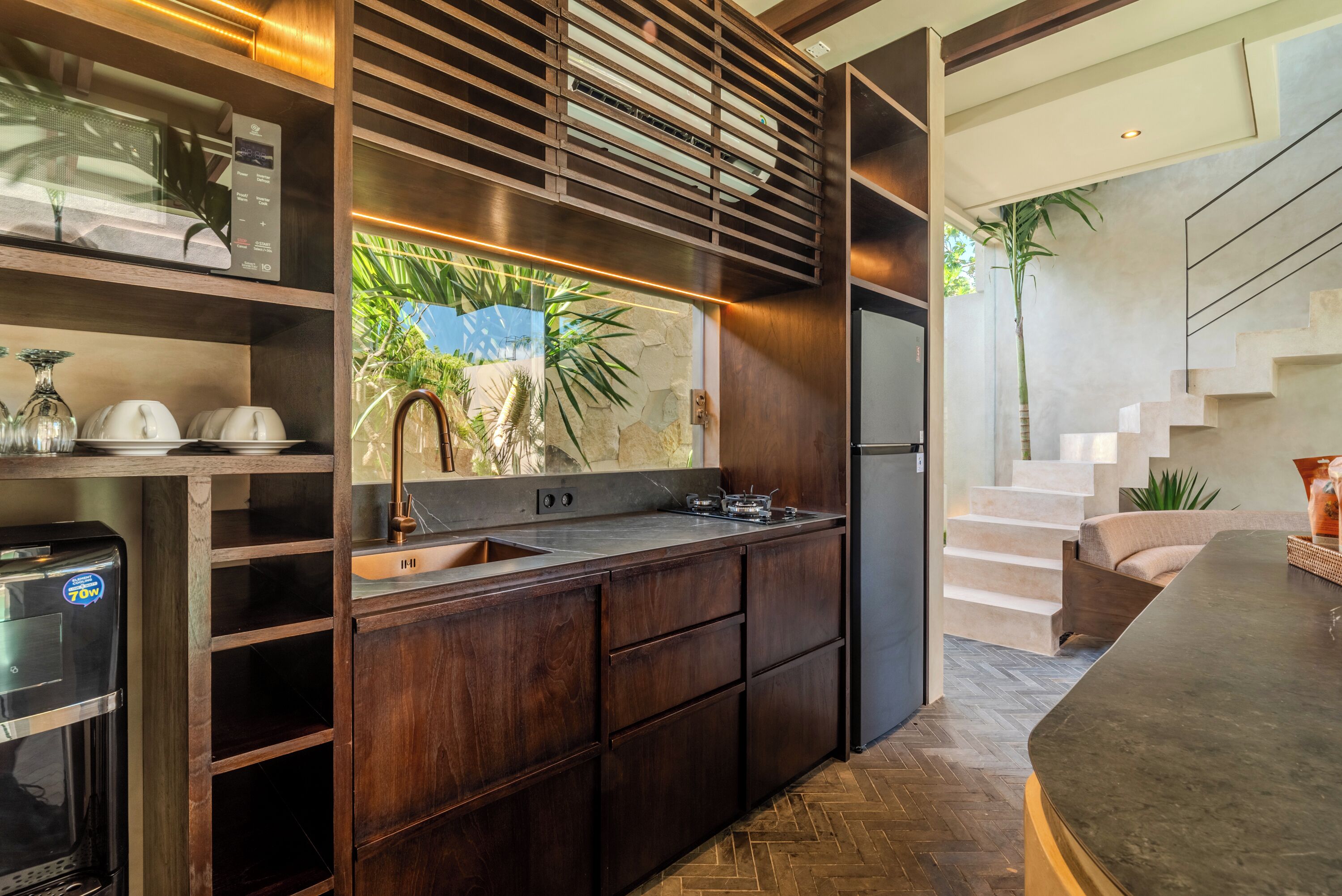 A modern kitchen with dark wood cabinets, a stainless steel sink, and a built-in stove. There is a large window with a view of palm trees and a stone wall.