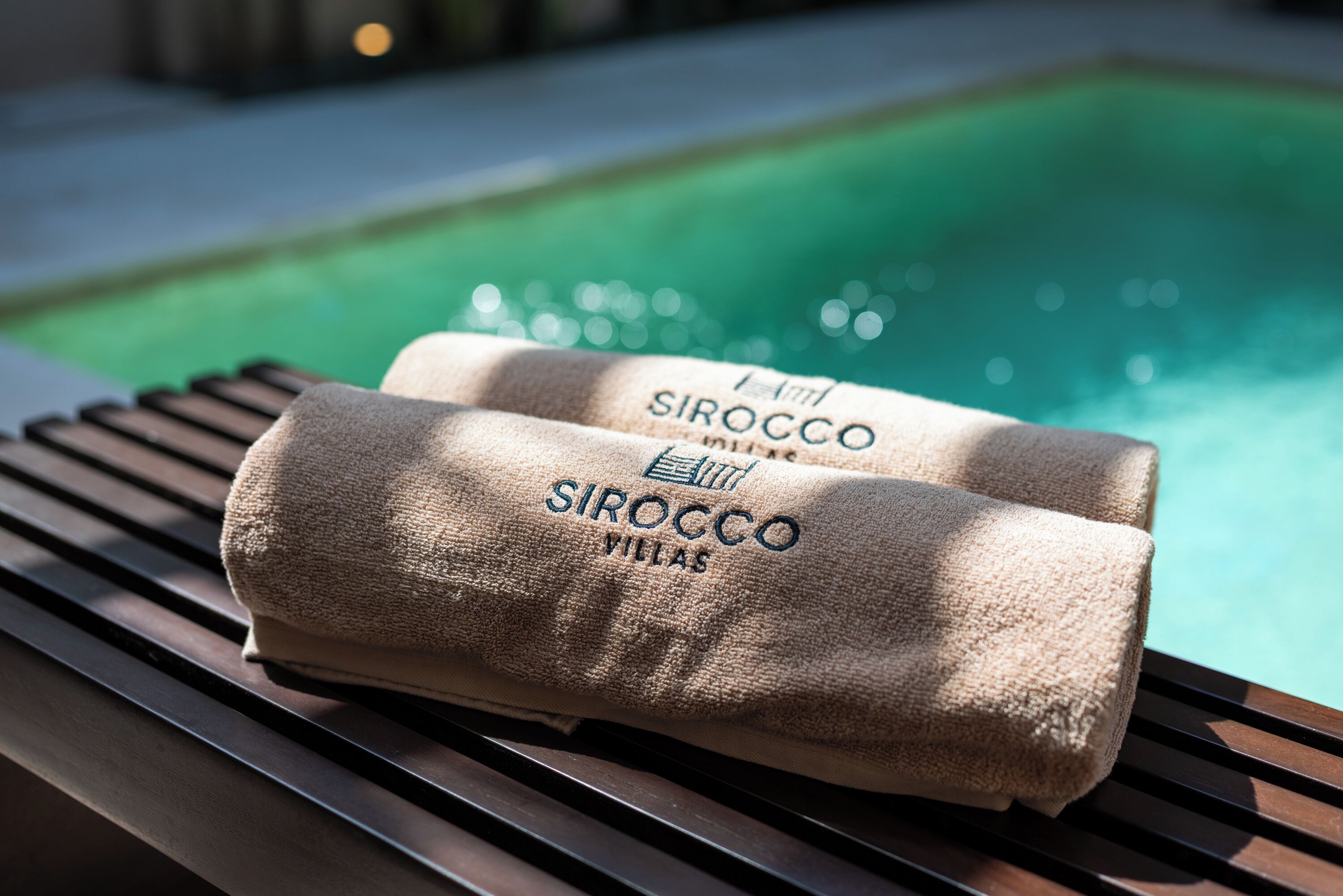 Two towels with "SIROCCO VILLAS" text on a wooden bench by a pool.