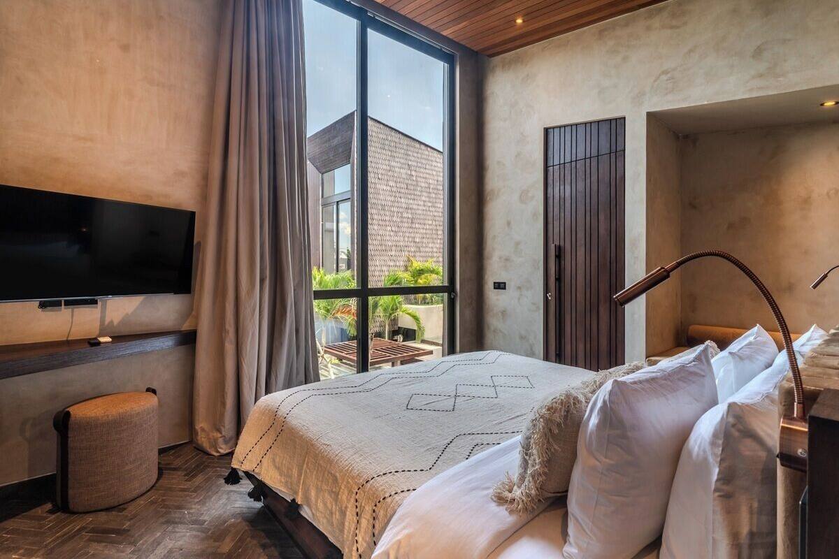Une chambre moderne dotée d’une grande fenêtre, d’un téléviseur à écran plat fixé au mur et d’un lit avec une literie blanche.