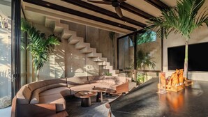 A modern living room with a curved sofa, a coffee table, and a fireplace. There is a staircase leading to an upper level, and a large window providing natural light. - Sirocco Villas Pererenan (Canggu)